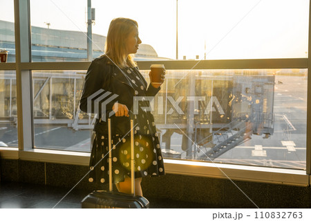 A young woman, holding a cup of coffee is ready for boarding with her suitcase. 110832763