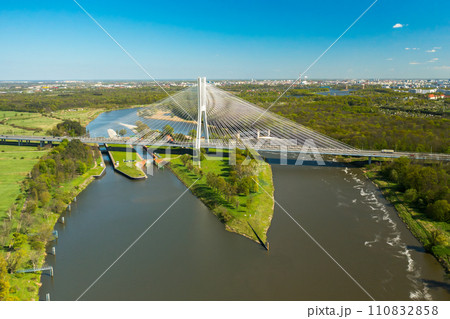 Cars drive on cable-stayed Redzinski Bridge over river flowing near scenic Wroclaw. Pylon bridge surrounded by lush green forests aerial motion along bridge 110832858