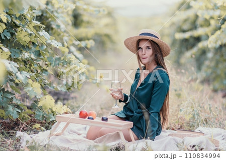 Woman picnic vineyard. Happy woman with a glass of wine at a picnic in the vineyard, wine tasting at sunset and open nature in the summer. Romantic dinner, fruit and wine. 110834994
