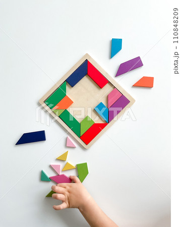 childs hand collects multicolored wooden mosaic on white background. child solves colorful tangram 110848209