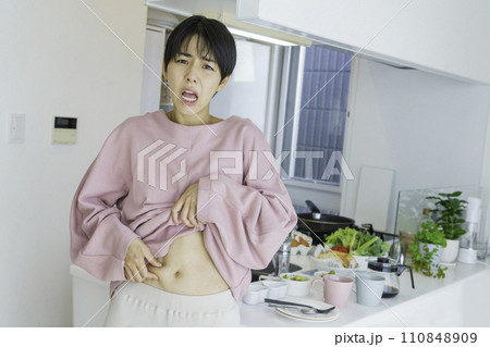 キッチンの前でお腹のお肉をつまんで贅肉を気にする若い女性 キッチンの前でお腹のお肉をつまんで贅肉を気にする若い女性 110848909