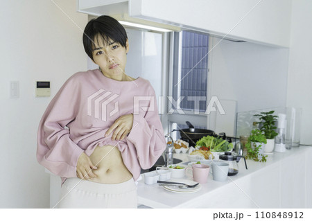 キッチンの前でお腹のお肉をつまんで贅肉を気にする若い女性 キッチンの前でお腹のお肉をつまんで贅肉を気にする若い女性 110848912