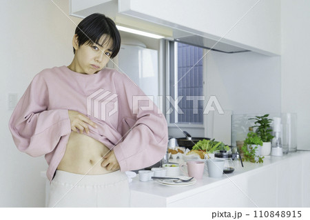 キッチンの前でお腹のお肉をつまんで贅肉を気にする若い女性 110848915