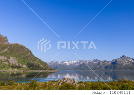 Lofoten's tranquil fjord mirrors rugged, snow-capped mountains under a clear azure sky 110849111
