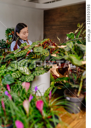 Vertical attractive and smiling young asian busy businesswoman, owner of flower shop. Spraying water and fertilizing herbs in pots. 110850398