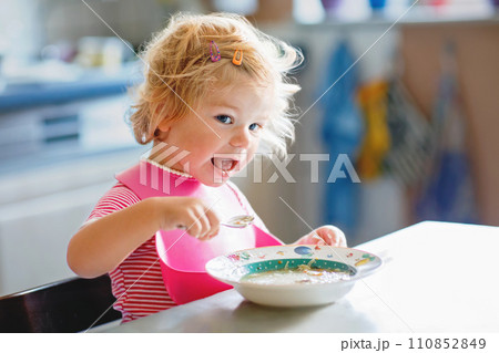 Adorable baby girl eating from spoon vegetable noodle soup. Healthy food, child, feeding and development concept. Cute toddler child with spoon sitting in highchair and learning to eat by itself Adorable baby girl eating from spoon vegetable noodle soup. Healthy food, child, feeding and development concept. Cute toddler child with spoon sitting in highchair and learning to eat by itself 110852849