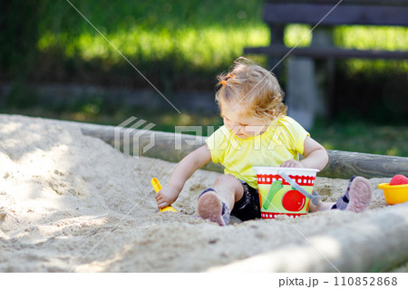 Cute toddler girl playing in sand on outdoor playground. Beautiful baby having fun on sunny warm summer sunny day. Happy healthy child with sand toys and in colorful fashion clothes. 110852868