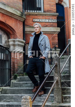 Handsome male model with gray hair in a black roll neck jacket and trousers, checked coat walking on the old city street. Cold season fashion stylish outfit trend. Old money aesthetic. Soft focus Handsome male model with gray hair in a black roll neck jacket and trousers, checked coat walking on the old city street. Cold season fashion stylish outfit trend. Old money aesthetic. Soft focus 110855491
