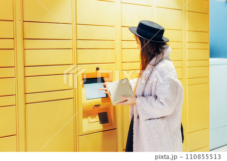 Fashion woman with box using modern postal automatic mail terminal with self service device for pickup or refund an order. Electronic locker for storing parcels. Online shopping. Selective focus. 110855513
