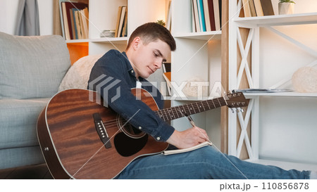 Song writing. Music composer. Guitar leisure. Talented inspired young man artist with string instrument creating melody in journal in home living room. 110856878