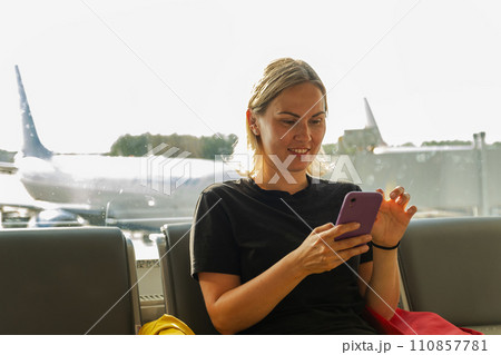 Airport terminal. Woman waiting for flight using smartphone. Girl with cell phone in airport surfing internet social media apps. Traveling female in boarding lounge of airline hub. Traveling girl 110857781
