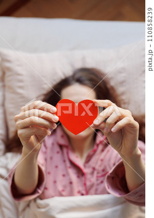 person in bed holds heart-shaped postcard in hands. surprise Valentine's day greeting from lover 110858239