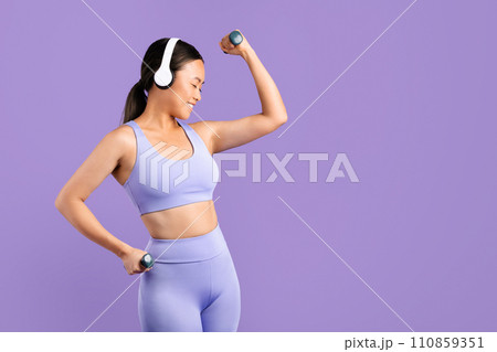 Asian woman in sportswear with headphones flexing muscles with dumbbells 110859351