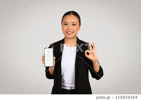 Content Asian businesswoman holding a smartphone with a blank screen in one hand and making an 'okay' 110859364