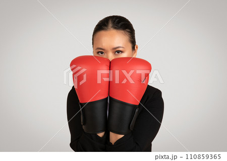 Determined Asian businesswoman with a focused gaze, peeking through red boxing gloves Determined Asian businesswoman with a focused gaze, peeking through red boxing gloves 110859365