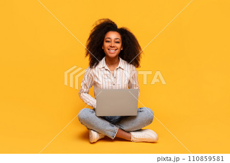 Content black woman with laptop sitting cross-legged on yellow backdrop 110859581