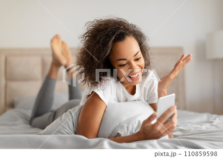 Joyful Black Millennial Lady Communicating Via Cellphone In Bed Indoors 110859598