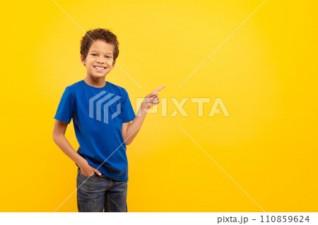 Cheerful black boy in blue shirt pointing at free space on yellow background 110859624