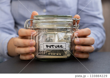 Unrecognizable woman holding Saving Money In Glass Jar filled with Dollars banknotes. FUTURE transcription in front of jar. Managing personal finances extra income for future insecurity 110864229