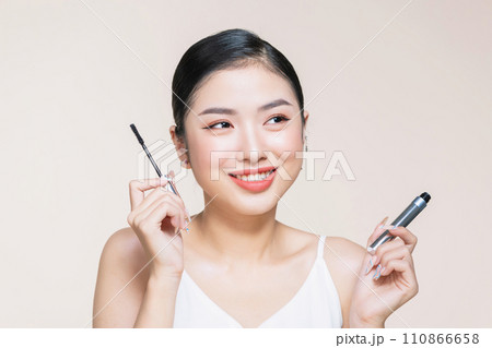 Beautiful asian young woman holding mascara and smiling on purple studio background, 110866658