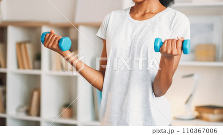 Dumbbells lifting. Home aerobics. Sporty slim woman in activewear practicing weights muscle domestic fitness training indoors in living room. 110867006