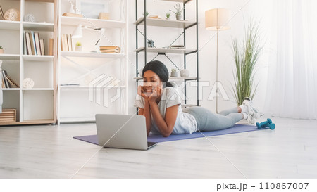 Laptop video call. Online workout. Internet communication. Joyful woman talking at virtual conference on fitness mat after yoga exercising in home living room. 110867007