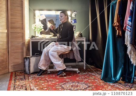 Full length portrait of female mime performer sitting by mirror backstage preparing for show in theater, copy space 110870123