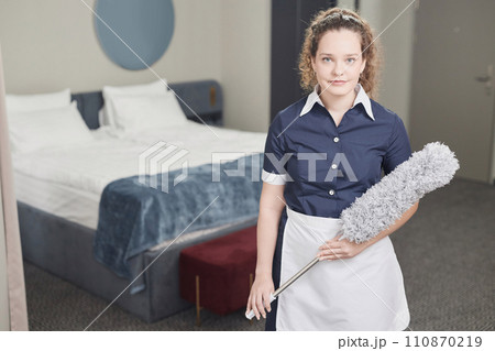 Waist up portrait of young woman as housekeeper wearing uniform and looking at camera holding duster in hotel room, copy space Waist up portrait of young woman as housekeeper wearing uniform and looking at camera holding duster in hotel room, copy space 110870219