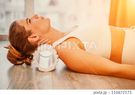 Adult athletic woman, in white bodysuit, performing fascia exercises on the floor - caucasian woman using a massage foam roller - a tool to relieve tension in the back and relieve muscle pain - the 110872573
