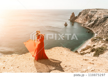 Woman red dress sea. Female dancer in a long red dress posing on a beach with rocks on sunny day. Girl on the nature on blue sky background. Woman red dress sea. Female dancer in a long red dress posing on a beach with rocks on sunny day. Girl on the nature on blue sky background. 110873297