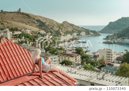 Woman sits on rooftop, enjoys town view and sea mountains. Peaceful rooftop relaxation. Below her, there is a town with several boats visible in the water. Rooftop vantage point. 110873340