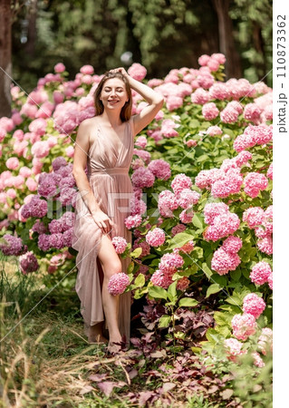 Hydrangeas Happy woman in pink dress amid hydrangeas. Large pink hydrangea caps surround woman. Sunny outdoor setting. Showcasing happy woman amid hydrangea bloom. Hydrangeas Happy woman in pink dress amid hydrangeas. Large pink hydrangea caps surround woman. Sunny outdoor setting. Showcasing happy woman amid hydrangea bloom. 110873362