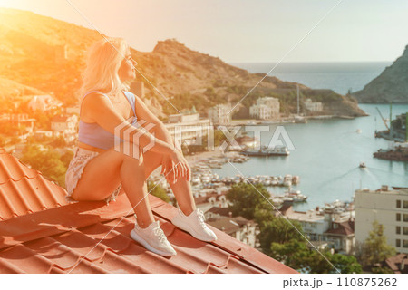Woman sits on rooftop, enjoys town view and sea mountains. Peaceful rooftop relaxation. Below her, there is a town with several boats visible in the water. Rooftop vantage point. 110875262