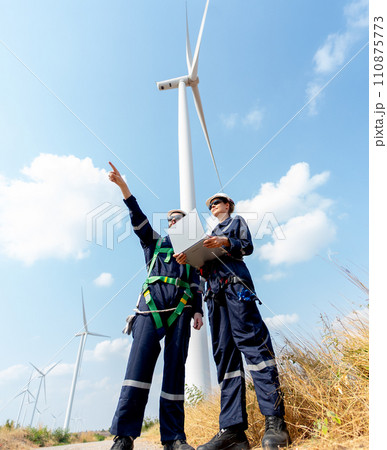 Vertical image of professional worker woman hold laptop and stand with her co-worker man point forward and they stand in front of wind turbine or windmill in the field for power plant business. Vertical image of professional worker woman hold laptop and stand with her co-worker man point forward and they stand in front of wind turbine or windmill in the field for power plant business. 110875773