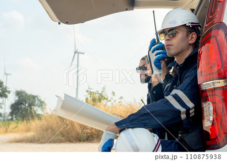 Close up technician worker man use walkie talkie to contact his co-worker and sit in the back of van in area of power plant with wind tubine or windmill. 110875938