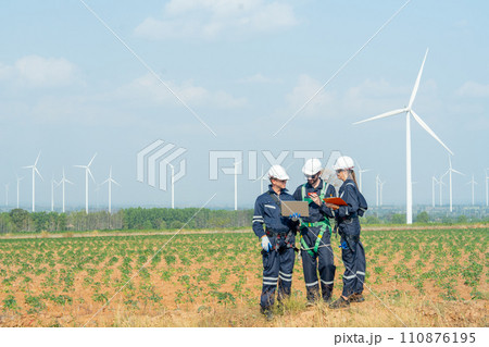 Side view technician workers man and woman stand in front of row of windmill or wind turbine and work together using laptop with day light. Side view technician workers man and woman stand in front of row of windmill or wind turbine and work together using laptop with day light. 110876195