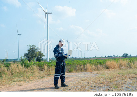 Wide shot of technician worker hold laptop and look to the monitor for working in front of windmill or wind turbine with blue skay in area of power plant factory. Wide shot of technician worker hold laptop and look to the monitor for working in front of windmill or wind turbine with blue skay in area of power plant factory. 110876198