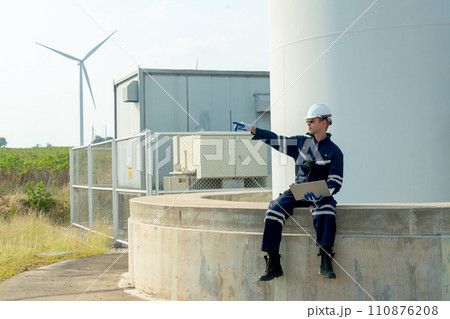 Professional technician worker hold laptop and sit on base of windmill or wind turbine also point to left side in area of green power plant. 110876208