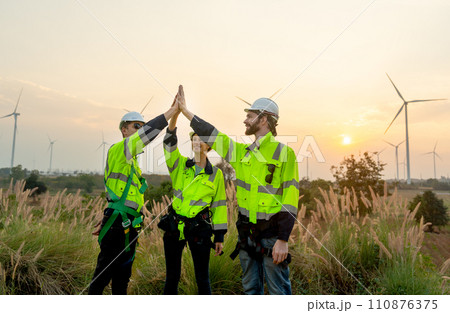 Group of technician worker man and woman stand with touch hands together express happiness during work and stand in front of windmill or wind turbine farm in area of power plant. 110876375