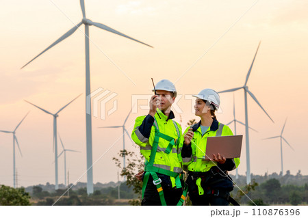 Professional technician man use walkie talkie to contact his co-worker and woman hold the laptop stand beside and work in front of windmill or wind turbine farm. 110876396