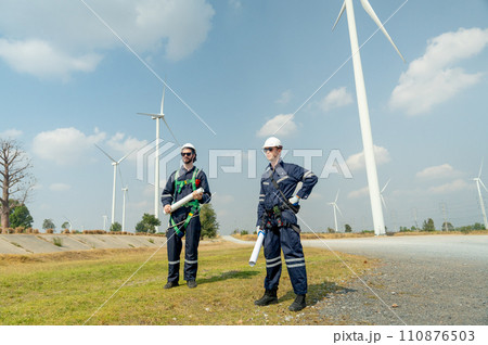 Professional Caucasian workers or technician with safety suit hold construction paper plan and stand in front of wind turbine or windmill in the row as power plant business. Professional Caucasian workers or technician with safety suit hold construction paper plan and stand in front of wind turbine or windmill in the row as power plant business. 110876503