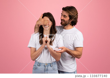 Happy young caucasian man covering woman eyes, hold gift box with red ribbon Happy young caucasian man covering woman eyes, hold gift box with red ribbon 110877140