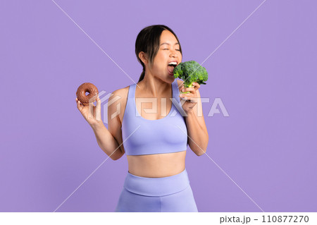 Asian woman in sportswear bites broccoli and holding donut Asian woman in sportswear bites broccoli and holding donut 110877270