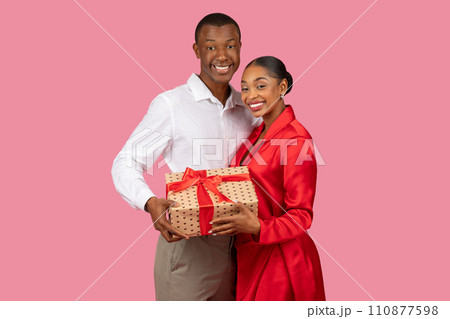 Black couple smiling with gift, man in white shirt, woman in red, pink background 110877598