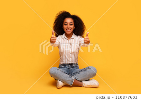 Happy black woman giving thumbs up while sitting on yellow backdrop Happy black woman giving thumbs up while sitting on yellow backdrop 110877603