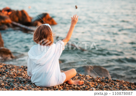 Summertime. Pre-school girl sitting alone on the beach and throws a pebble to the water. Back view. Ocean at the background. Concept of adoption and children's day Summertime. Pre-school girl sitting alone on the beach and throws a pebble to the water. Back view. Ocean at the background. Concept of adoption and children's day 110878087