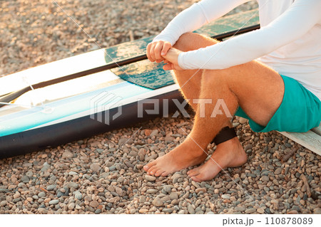 A man sitting at the pebble beach with a sup board. Feet and surfboard close up. Summer recreation and activity A man sitting at the pebble beach with a sup board. Feet and surfboard close up. Summer recreation and activity 110878089