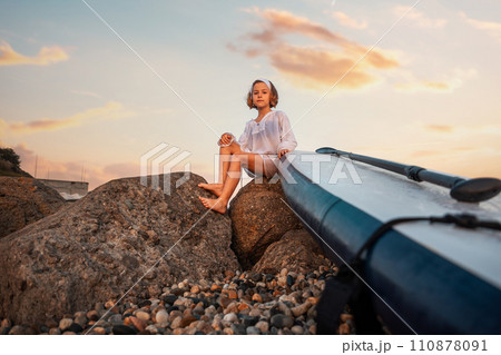 Summer activity vacations. Pre-school girl sitting with sup board at big beach rocks. Bottom view. Sunset sky at the background 110878091