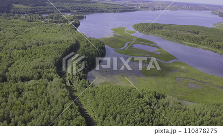 Aerial view over sunny lake surrounded by pine tree forest near town. Clip. During sunny summer day. Top view of the forest lake near the town 110878275