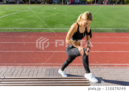 A fit woman pauses to look at her fitness tracker during a workout session on a sunny track field. 110879249